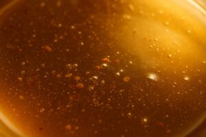 تصویر نزدیک از سطح عسل طبیعی طلایی با ذرات گرده گل و حباب‌های ریز در نور طبیعی